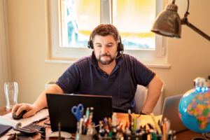 Man with headphones and microphone having conference call online sitting at home office with two laptops. Working from home during quarantine and self isolation period at pandemic. Manager at online meeting upset with markets falling down