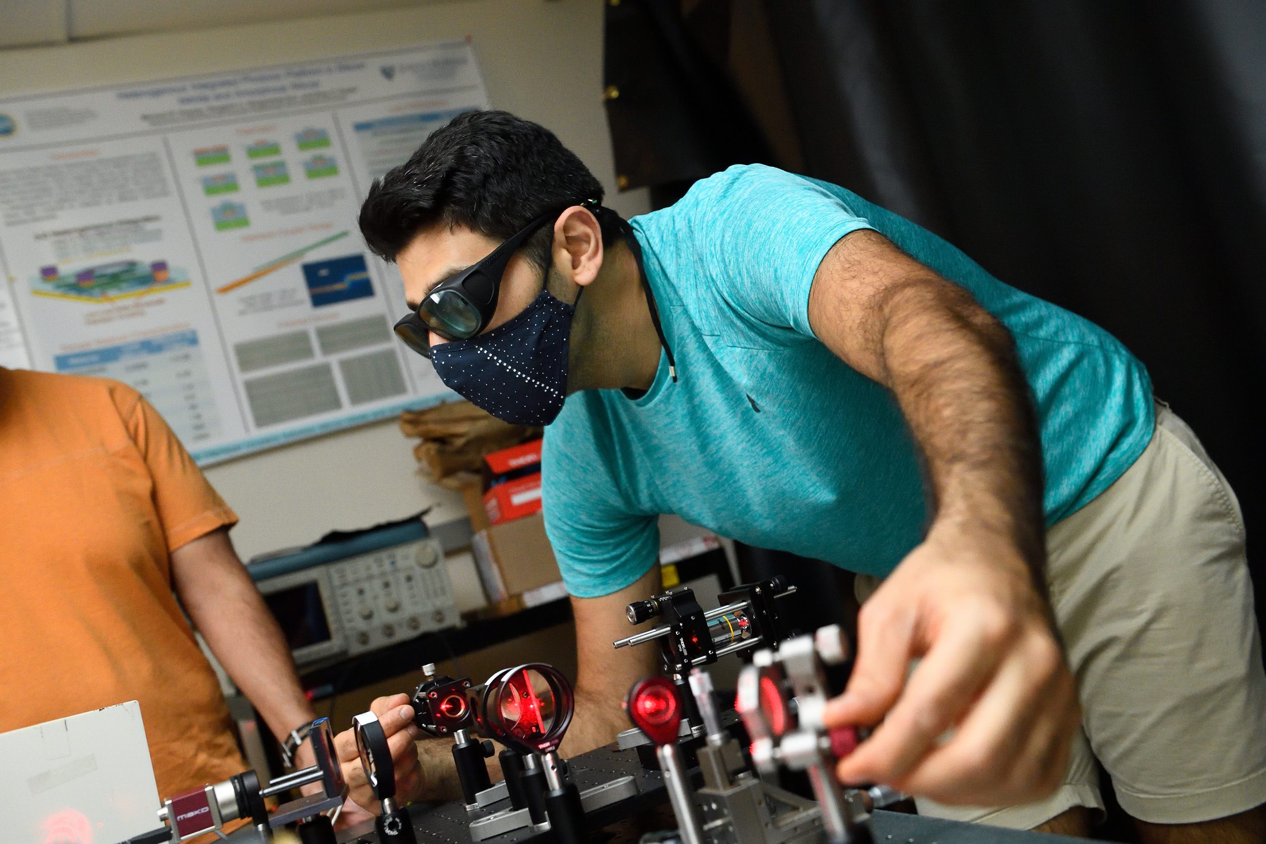 A male student wears goggles and a face mask while adjusting a piece of equipment.