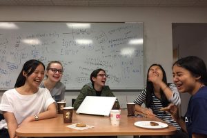 Five students sit around a table laughing, drinking coffee and eating cookies. A wipe board covered in equations and graphs is on the wall behind them.