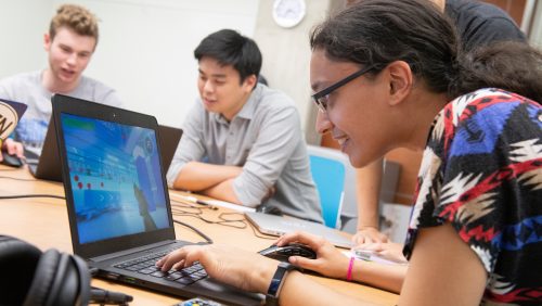 A student plays a video game on a laptop computer while another student watches over their shoulder. Two students in the background are also looking at another laptop screen.