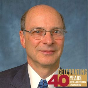 A headshot photograph of Ross Corotis wearing a suit and a blue tie against a blue backdrop. In the lower right corner is a graphic "Celebrating 40 years Civil and Systems Engineering.