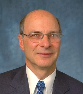 A headshot photograph of Ross Corotis wearing a suit and a blue tie against a blue backdrop.