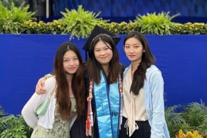 Three women posing for a photo. Yiran "Gigi" Wang is in the center in graduation attire of a cap and gown. 