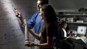 Two people discussing over a white board and woman is sitting on a chair with her monitor in the background.