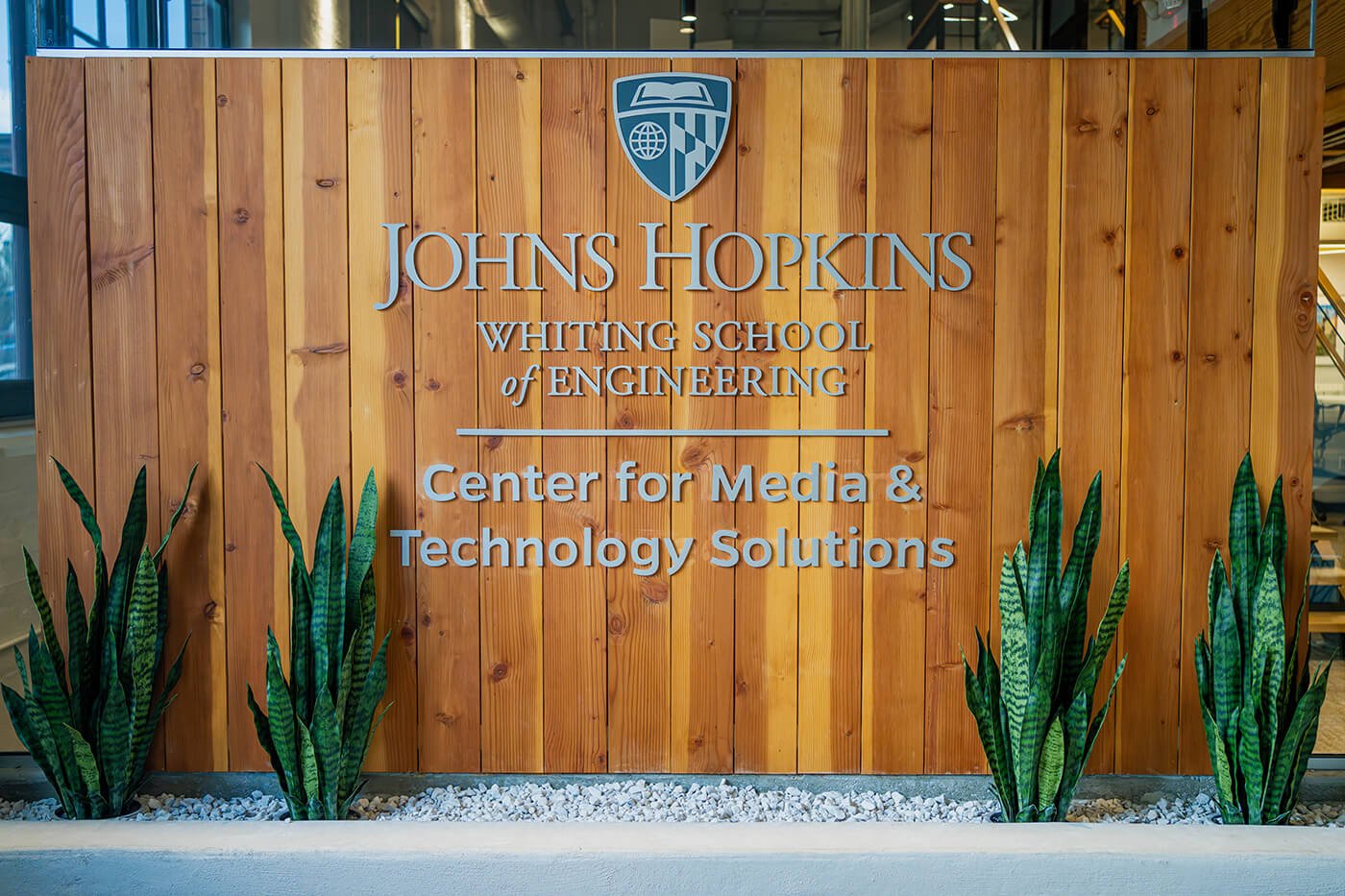 Stieff Suite 110 photo of lobby signage which is Johns Hopkins Whiting School of Engineering Center for Media and Technology Solutions on a wood lobby sign surrounded by snake plants.