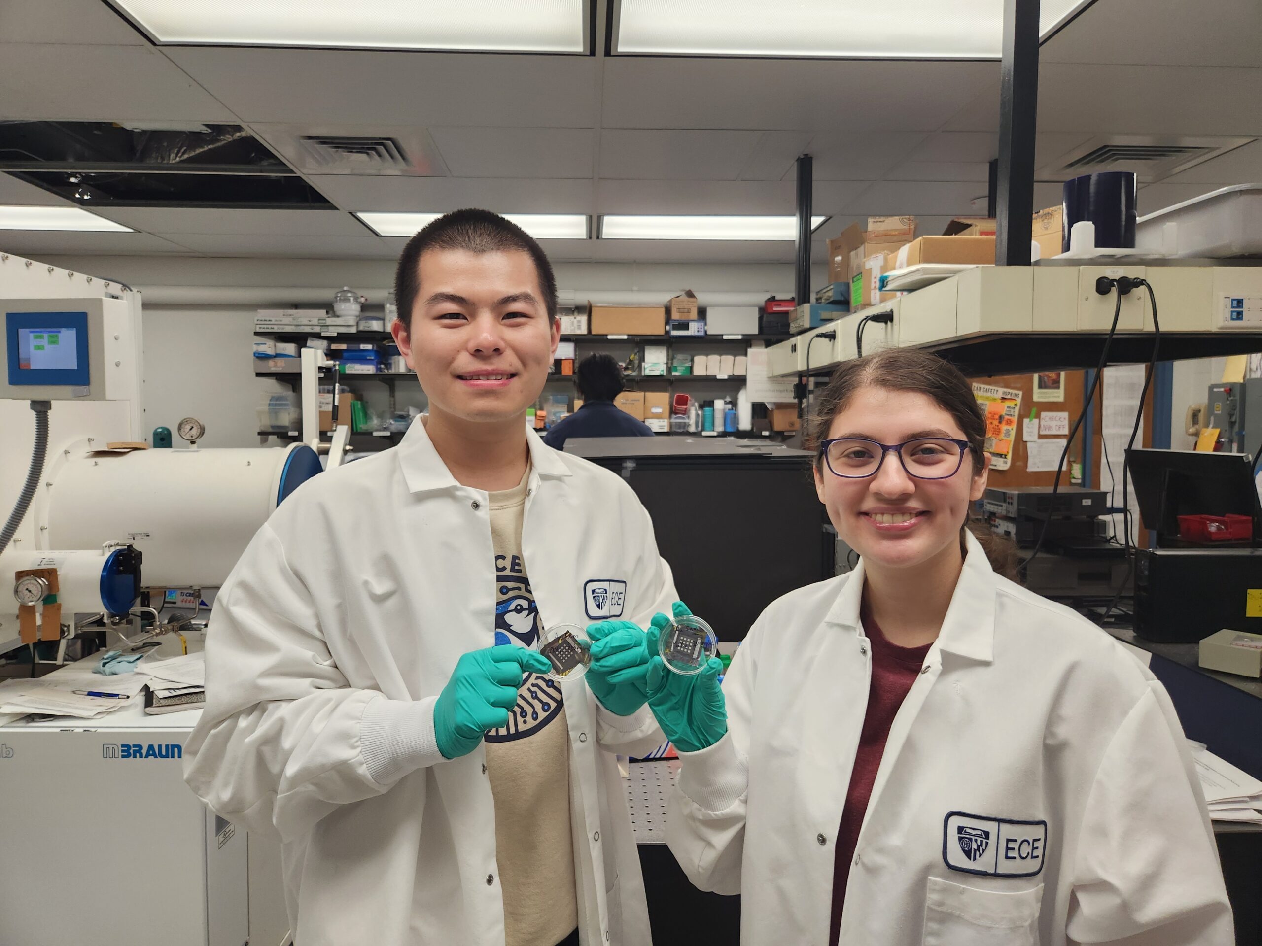 Two students with lab coats stand side by side in a research laboratory, each holding a small electrical device, carefully presenting the devices for inspection.