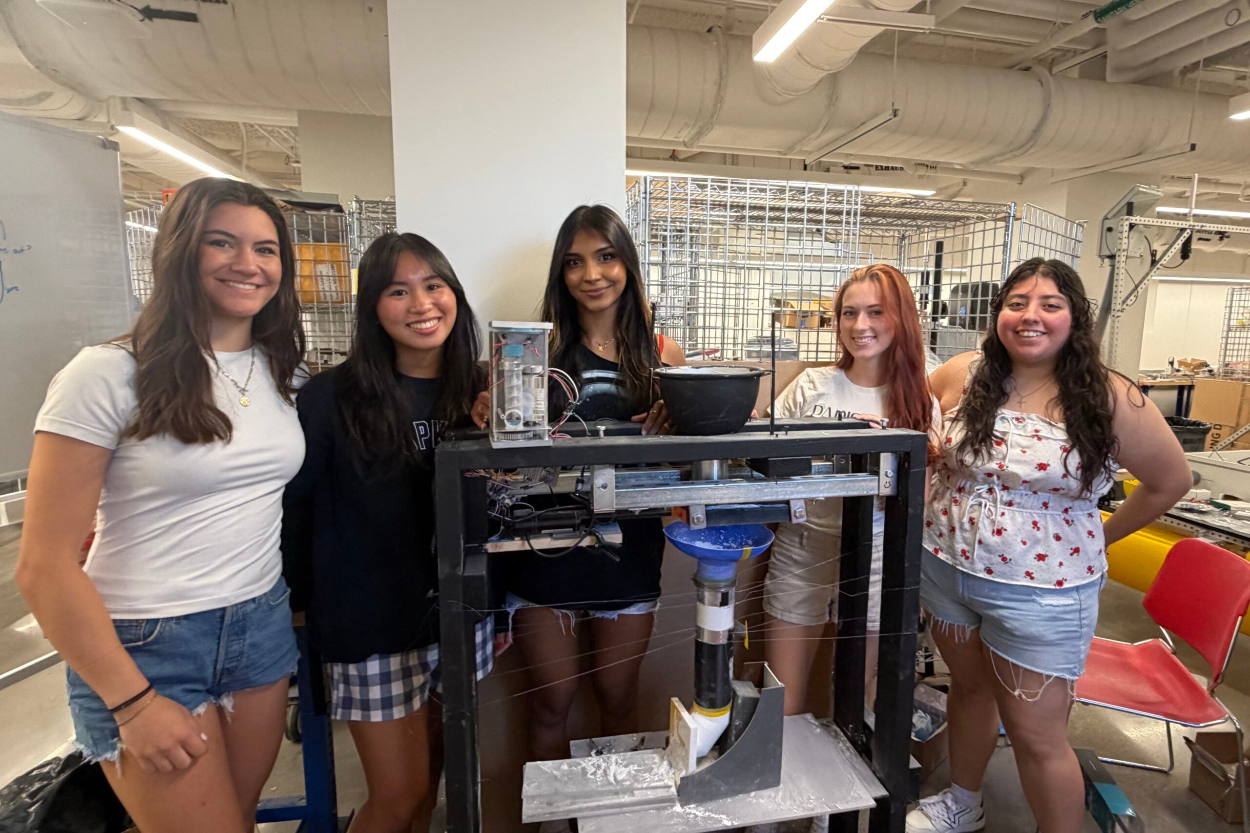 Five team members stand behind a wooden frame that holds their small prototype regolith auger
