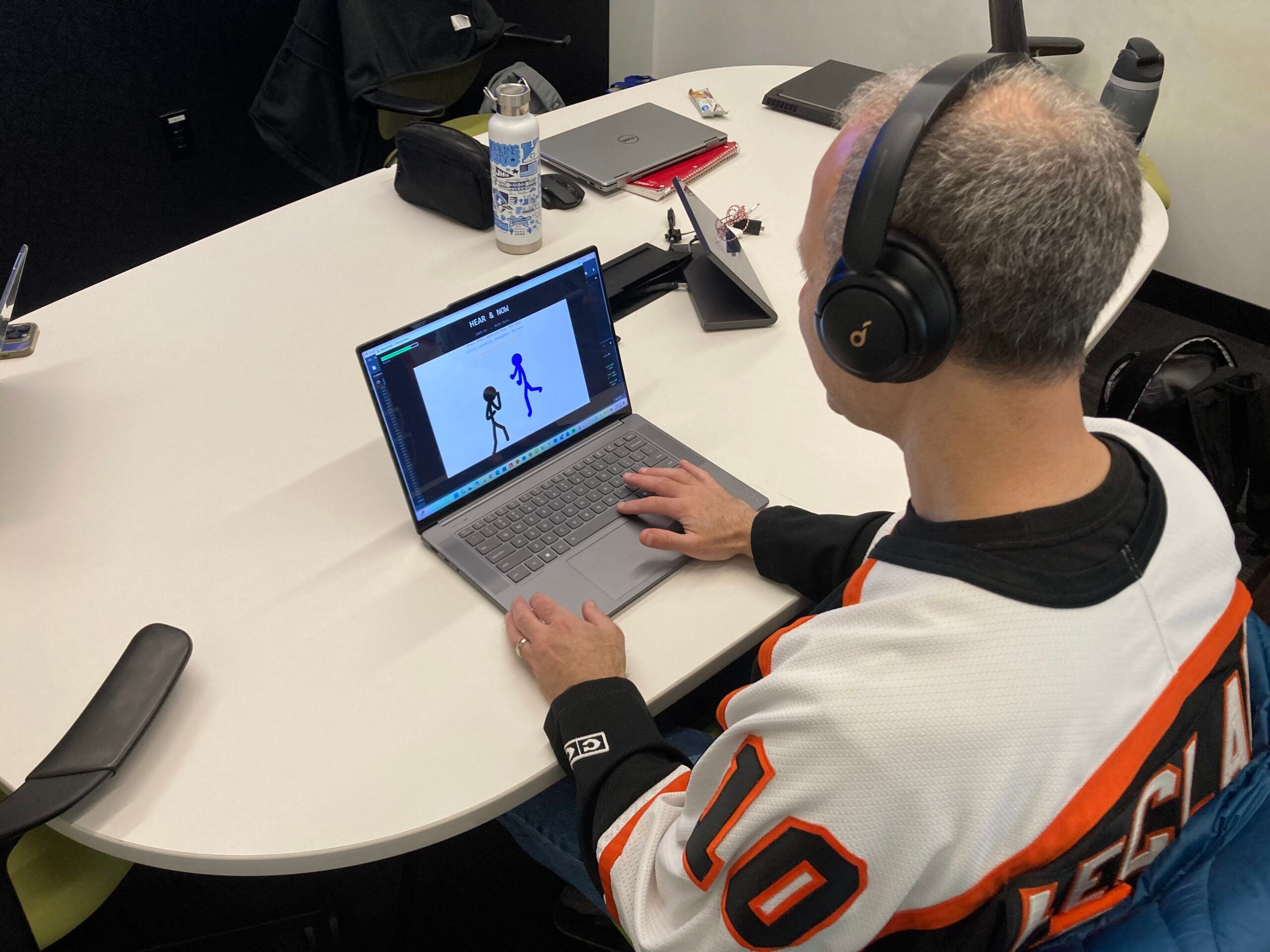 A person wearing headphones and a sports jersey sits at a white table, viewing a laptop screen displaying the Hear & Now application. The screen shows two stick figure animations in a blue-tinted interface. A water bottle, laptop bag, and second laptop are visible in the background.