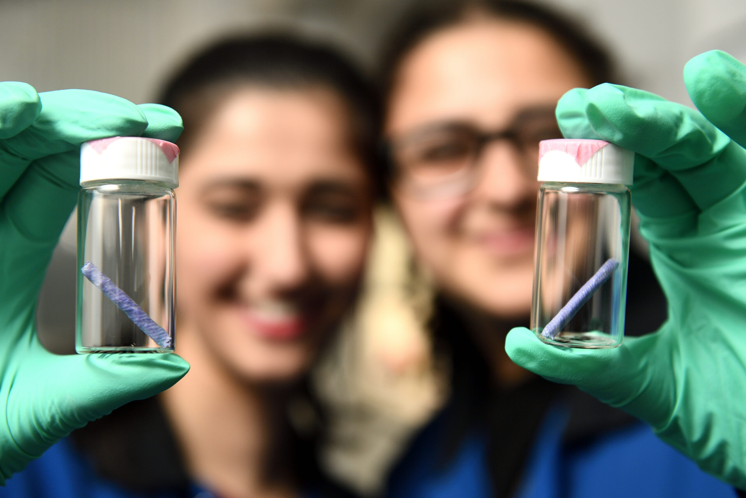 Two researchers in green gloves hold up glass vials each containing a small purple biodegradable stent scaffold.
