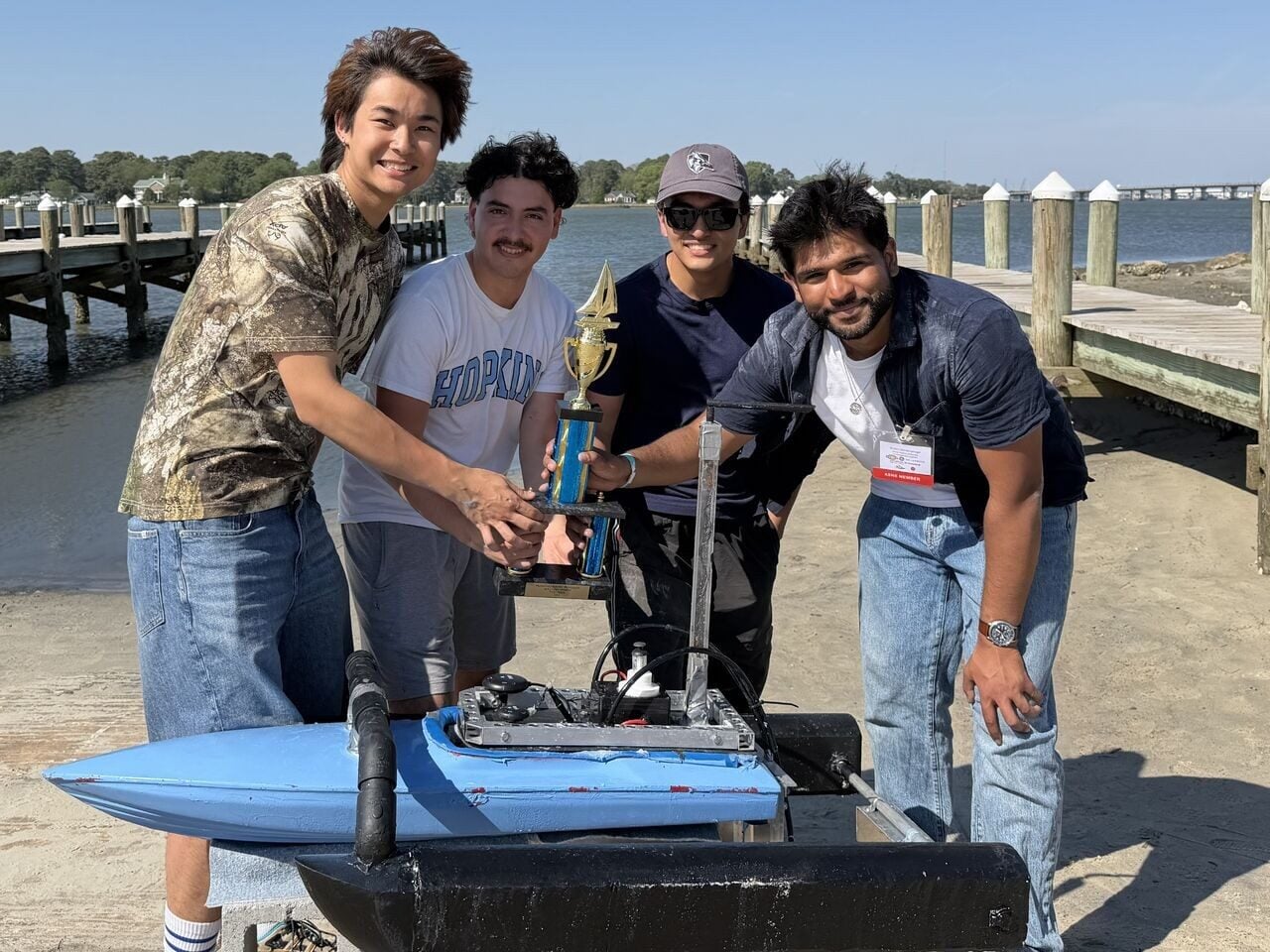 Four members of the team stand around a small blue and black RC boat and hold a trophy