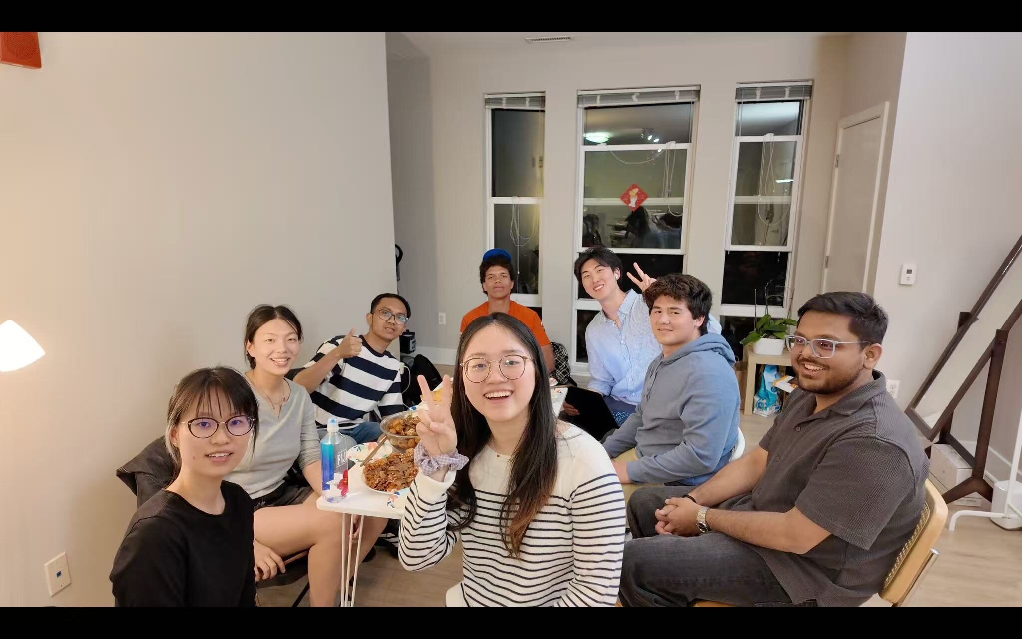 A group of undergraduate and graduate students from diverse backgrounds sit together indoors after a Flavor Journey cooking event, smiling and sharing food in a warm, social setting.
