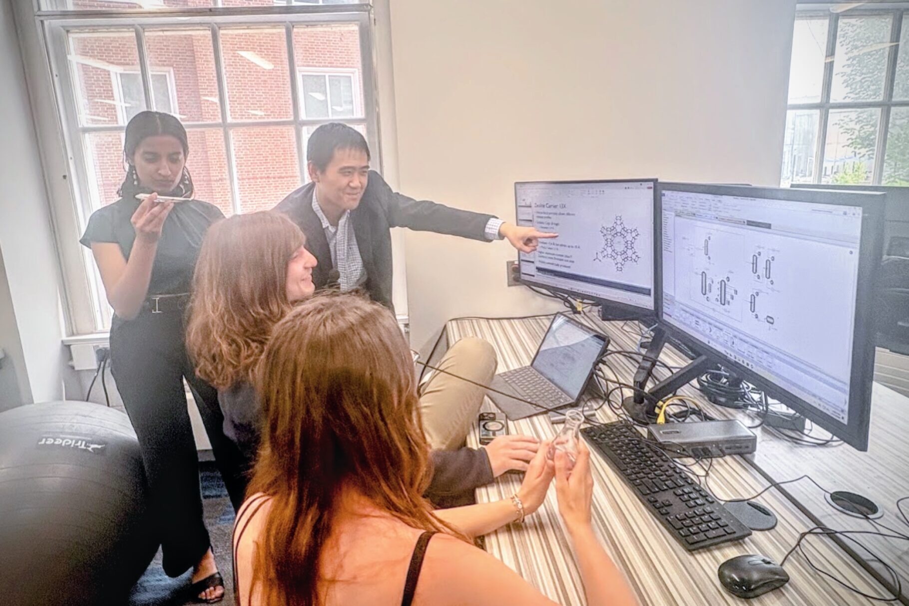 Ze[r]osion team members discuss zeolite molecular structures and an ASPEN process flow diagram displayed on two monitors that they are clustered around