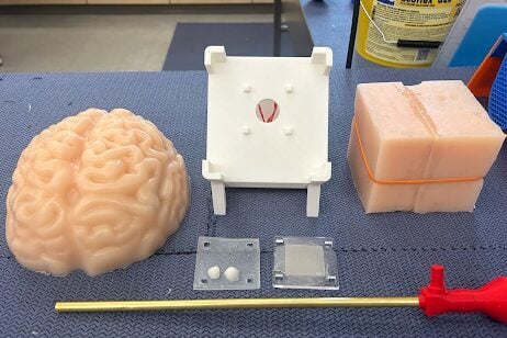 This image shows the disassembled simulator components: a silicone brain cap, a white stand, two clear square sheets, a mimetic endoscope, and silicone block.
