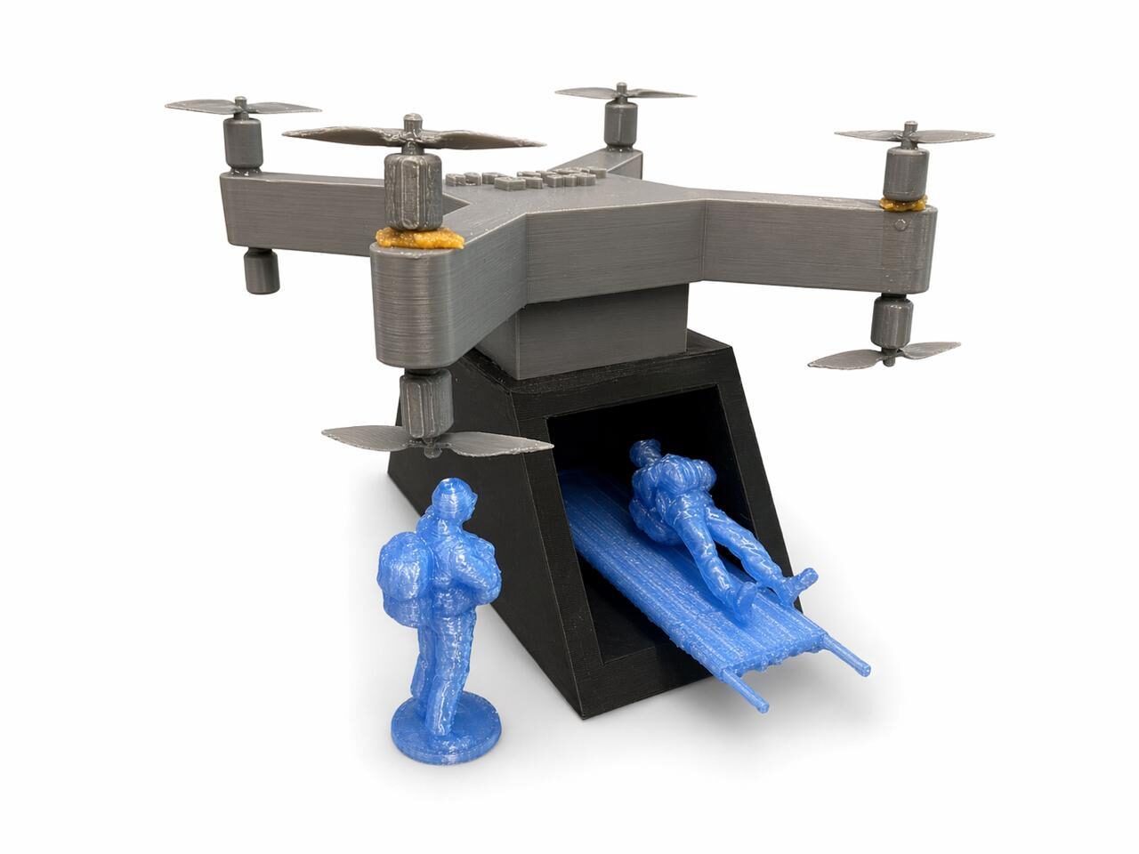 A small 3D-printed model of a drone with an open rear carriage is shown. A miniature figure lies on a stretcher being loaded into the drone, while another figure stands nearby, demonstrating the casualty evacuation use case.