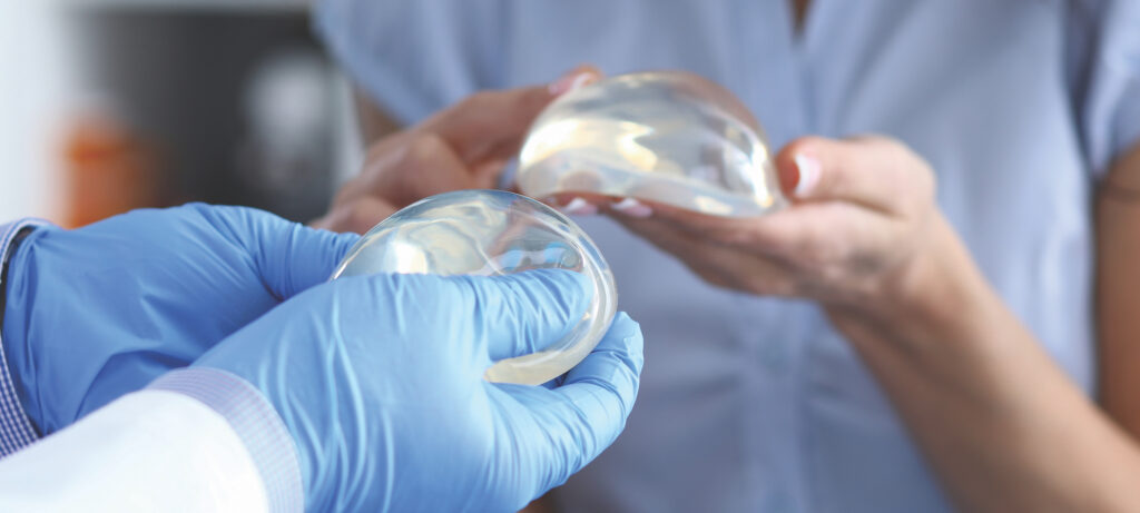 Two people—one wearing a doctor's white coat and the other wearing scrubs—hold silicone breast implants in their hands.