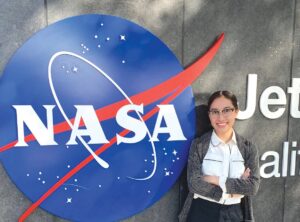 Photograph of Katya Echazarreta, smiling with arms crossed in front of her body, standing in front of a NASA sign