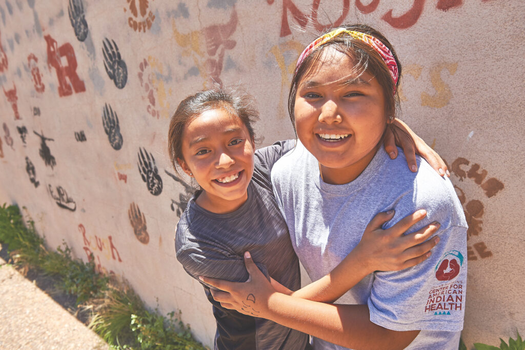 Two teenage girls from the White Mountain Apache Tribe embrace each other.