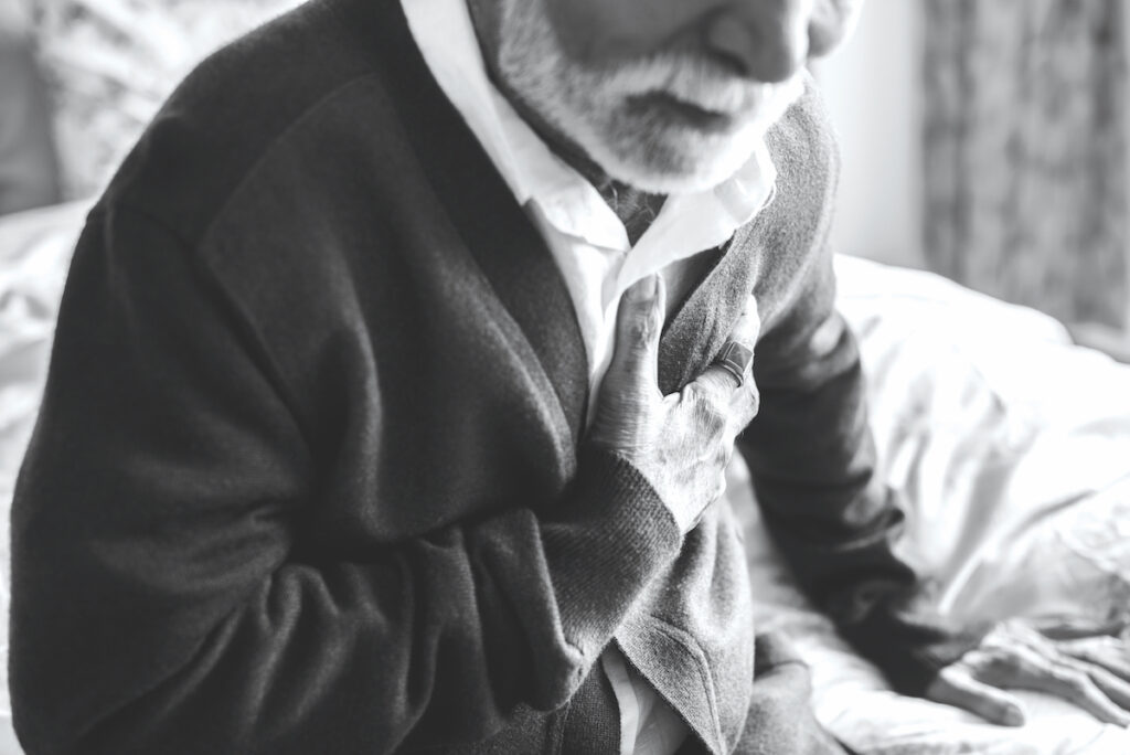 An elderly man in a sweater vest clutches his chest in pain