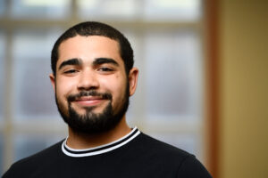 Photo of Allan Raposo smiling, standing in front of a window