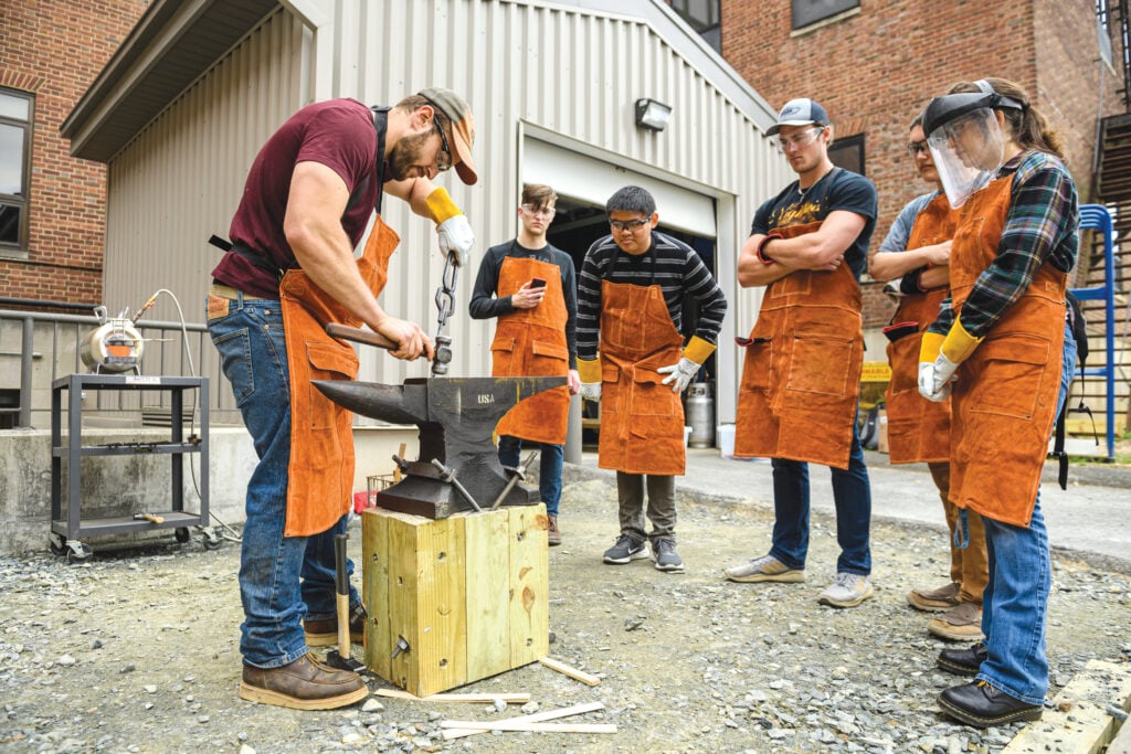 Photograph of group of students watching one student shape softened metal on an anvil with a hammer.