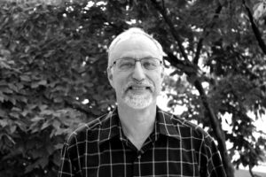 Howard Katz black and white photo of him smiling in front of trees with plaid shirt.
