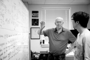 black-and-white image of Alan Yuille in present day as a professor, shown in front of a white board discussing its contents with a student