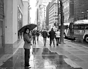 black-and-white image of a woman holding an umbrella on a rainy busy city street with passersby in the background