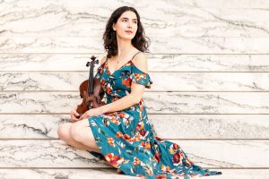 Joanna wears a flowing off the shoulder dress and sits on marble steps on Homewood campus holding her fiddle.