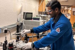 A smiling scientist in a blue lab coat and safety goggles works on equipment in a lab, conveying a sense of focus and innovation.