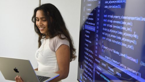 Smiling woman standing in front of a monitor with coding on it.