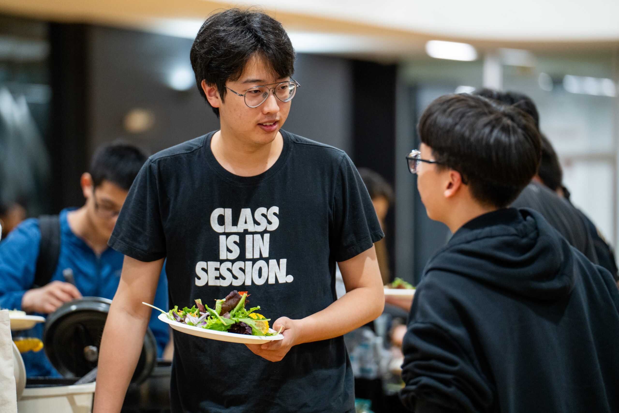 Students standing in line and talking while holding plates of food at the 2024 Masters graduate BBQ.