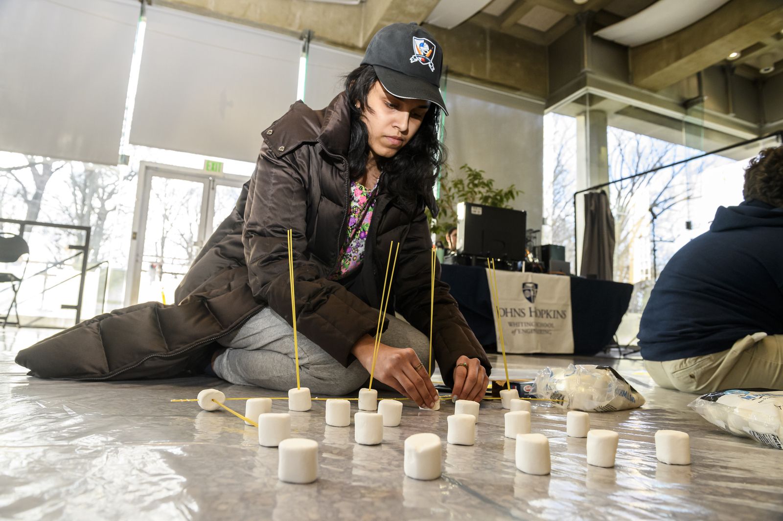 Student inserting dry spaghetti noodles into marshmallows spread on the floor.