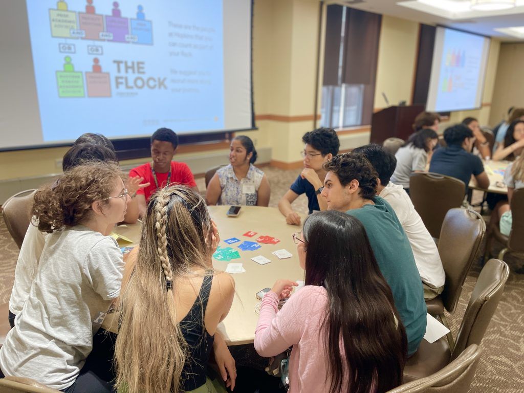 Students gathered at a table in from of a screen displaying a slide stating "Meet the Flock."