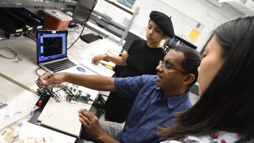 A professor who is explaining the breadboard circuit connection to two students in the laboratory.