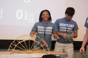 Two students wearing Hopkins Engineering Innovation t-shirts with spaghetti bridge on the desk.