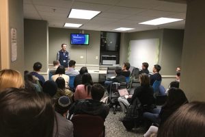 A man who is standing and speaking to a group of people who are sitting in a room.