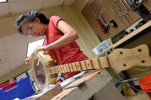 A woman building a wooden banjo instrument, while wearing protective eye gear.