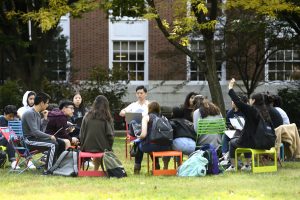 A group of students who are sitting on chairs in a garden area outdoors on a campus.