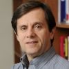 A headshot of Peter Kazanzides wearing a gray shirt with a bookshelf in the background