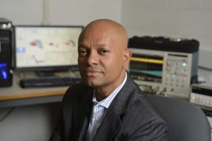 A headshot of Ralph Etienne-Cummings wearing a gray blazer while sitting on a chair with his workstation behind him.