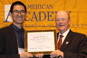 Qian Zhang receiving an award from a man with yellow banner in the background saying " American Academy".