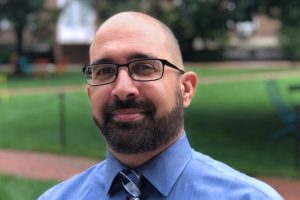 A headshot of Steven Marra wearing a blue shirt with a tie and standing outdoors of the campus.
