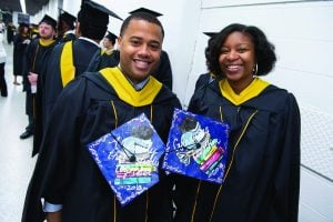 Two students in their graduation gown posing for a picture while holding their painted caps.