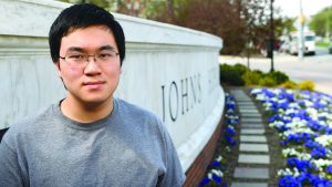 A headshot of a male student standing beside the "Johns Hopkins University" wall outdoors of the campus.