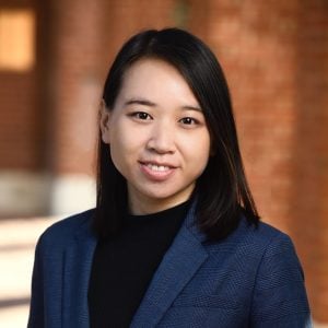 A headshot of Anqi (Angie) Liu wearing a dark blue blazer and standing in the corridors of the campus.