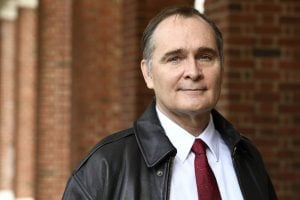 A headshot of James Bellingham wearing a leather jacket and standing in front of brick pillars in the background