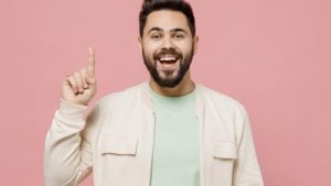 A man in a jacket pointing his right hand finger to the top with pink background.
