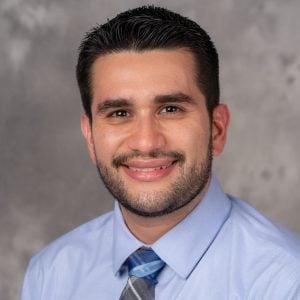 A headshot of Dr. Corey Oses who is against a plain background. He has short dark hair and is wearing a collared shirt and tie.