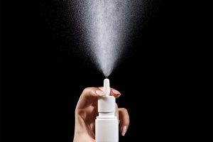 A hand activates a nasal spray bottle, spraying mist into the air.