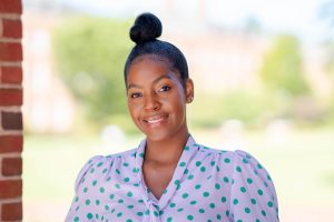 Head shot of Ayana Teal wearing lavender puff sleeve blouse with green polka dots with blurred greenery in background